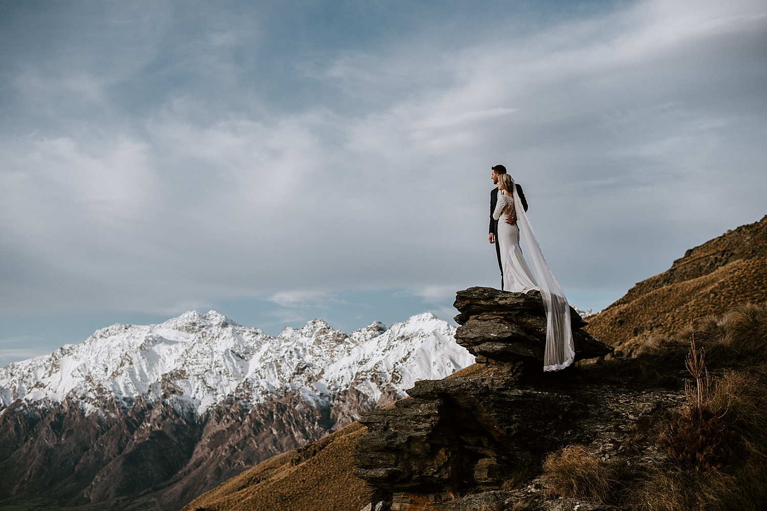 Newly wed couple embrace on a cliff in front of snow capped hills in Queenstown.