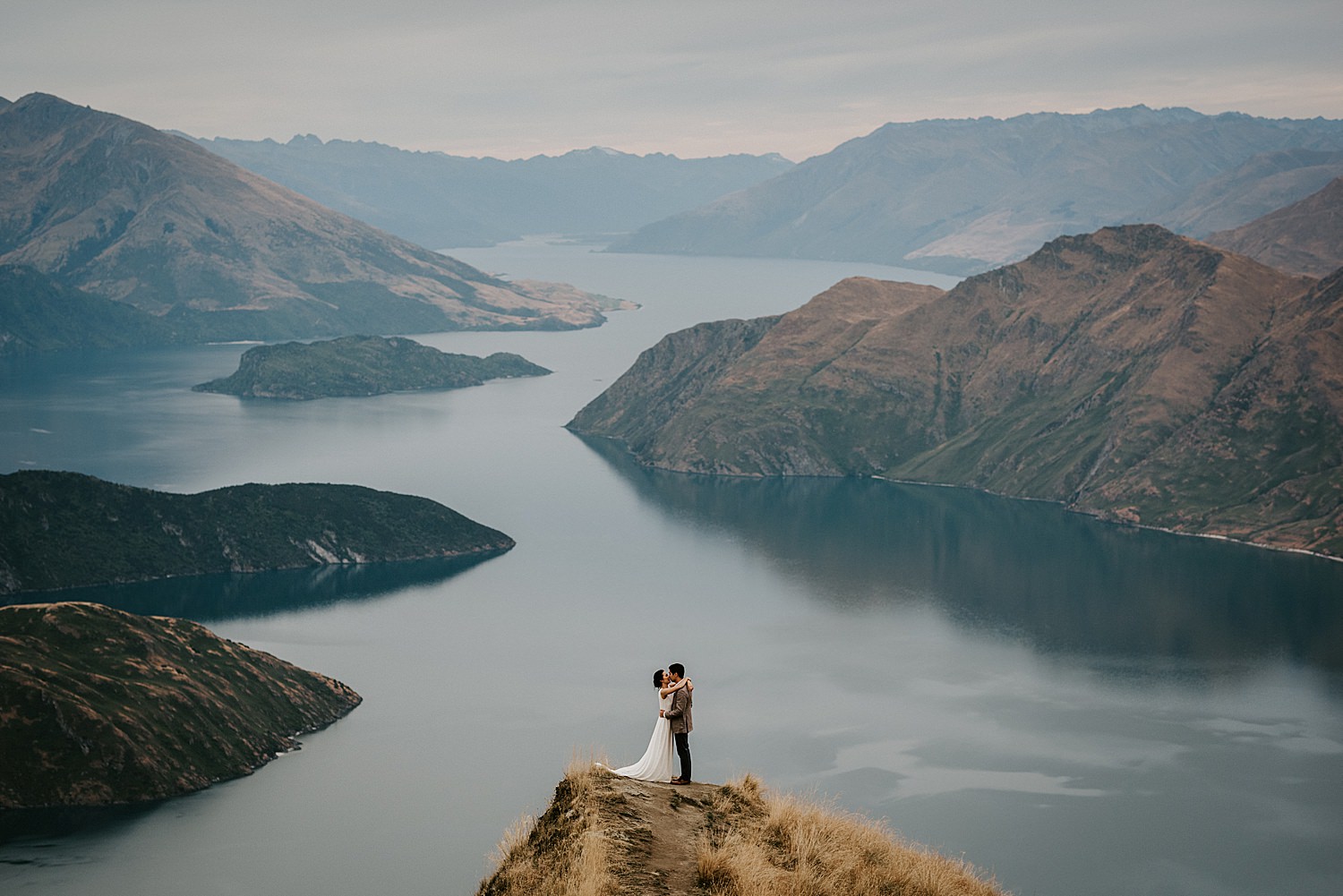 Couple embrace on a high peak with the lake and rolling mountains in the background near Queenstown.