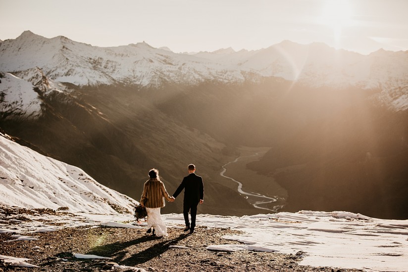 A couple walk hand in hand on snowcapped mountains, with a sunset in the background near Queenstown.