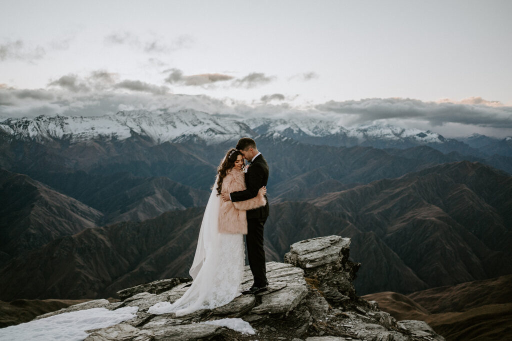 couple high above the mountaisn hugging into each others houlders in thier wedding outfits, with snow capped hills in the background