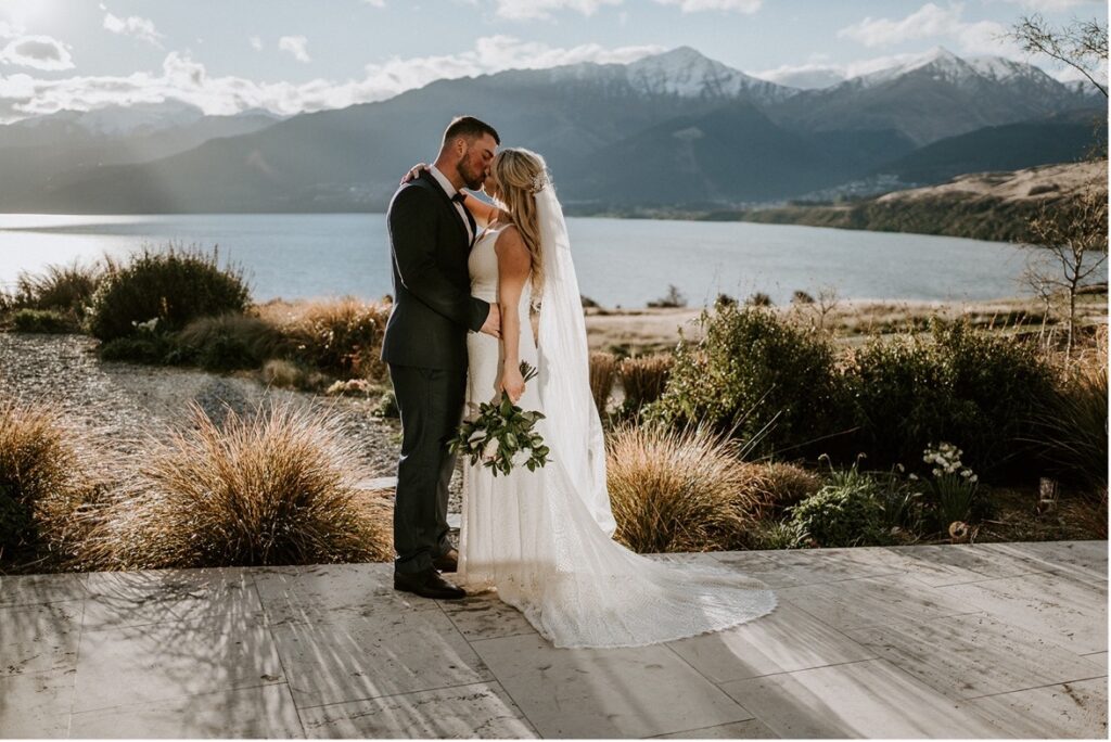 Newly wed couple standing on a platform kissing with the lake and mountain views in the background.